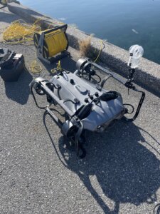 Remotely operated underwater vehicle on a concrete dock with a yellow tether spool and cables by the water.