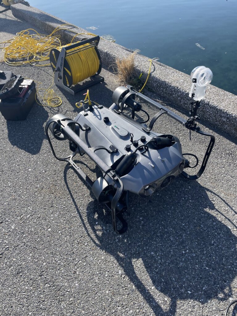 Remotely operated underwater vehicle on a concrete dock with a yellow tether spool and cables by the water.
