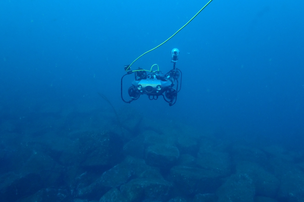 rov-underwater-5 - 株式会社SeaChallenge Remotely operated underwater vehicle (ROV) with thrusters and a trailing cable hovering above a rocky seabed in clear blue water