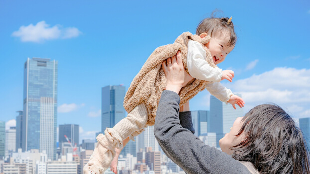 33490786_s - 株式会社SeaChallenge Parent lifting a smiling baby in the air against a city skyline on a sunny day.