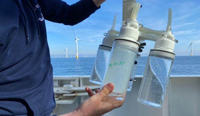 Researcher on a boat collecting seawater samples from a sampling rig, with offshore wind turbines in the background.