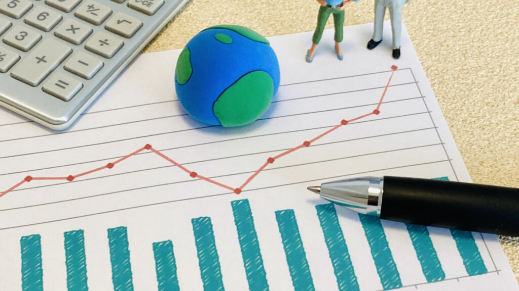 Desk scene with a blue-green clay globe, tiny figurines, and a red upward-trending line chart overlaid on a printed graph with blue bar chart; calculator and pen nearby.