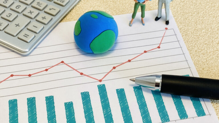 Desk scene with a blue-green clay globe, tiny figurines, and a red upward-trending line chart overlaid on a printed graph with blue bar chart; calculator and pen nearby.