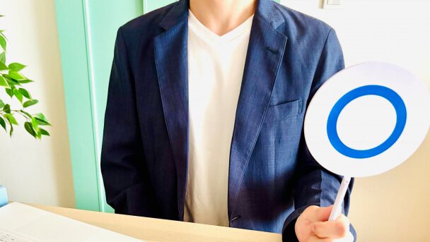 Person in a navy blazer and white shirt sits at a desk, holding a round sign with a blue ring.