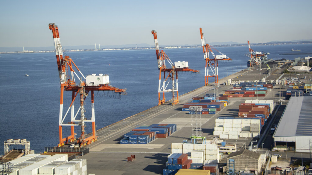 Port terminal with tall orange gantry cranes along a pier, blue sea to the left and stacked shipping containers in the foreground and background.