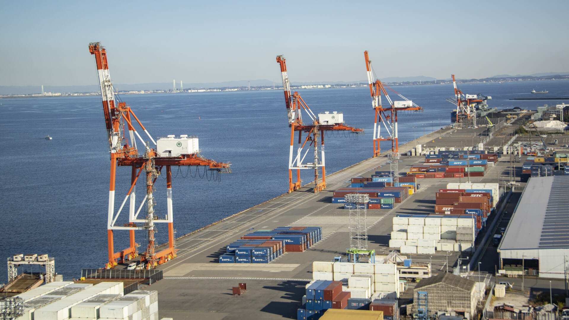 Port terminal with tall orange gantry cranes along a pier, blue sea to the left and stacked shipping containers in the foreground and background.