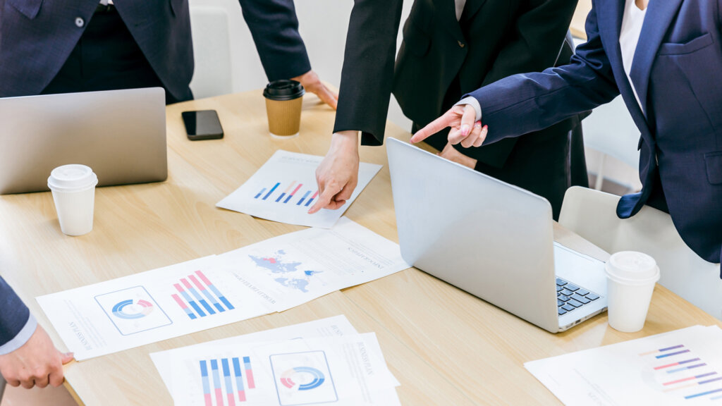 Group of professionals in suits leaning over a conference table, pointing at printed charts and a laptop.