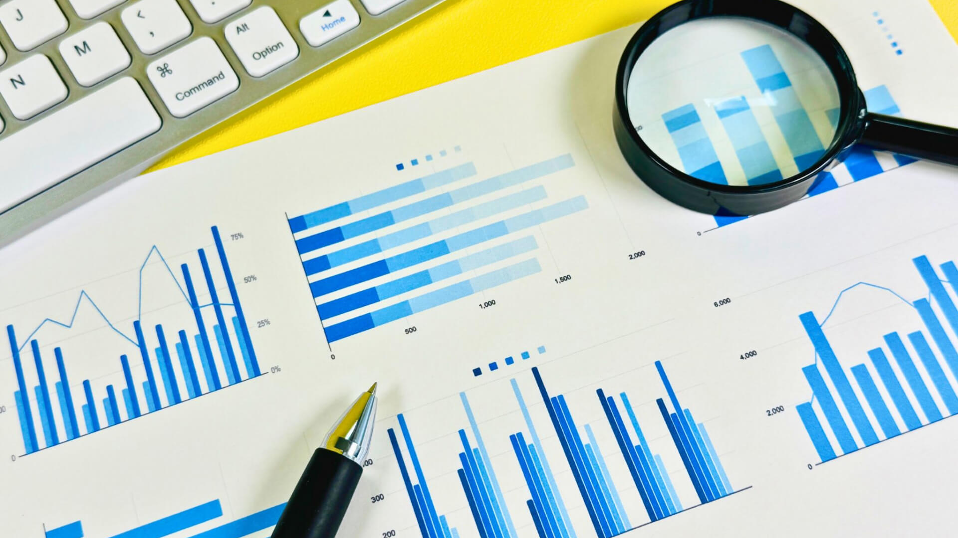 Blue bar and line charts on printed reports with a magnifying glass, keyboard, and pens, suggesting data analysis.
