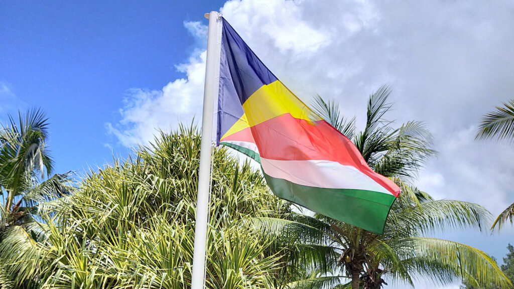 Seychelles flag fluttering on a white pole with palm trees and a blue sky behind.