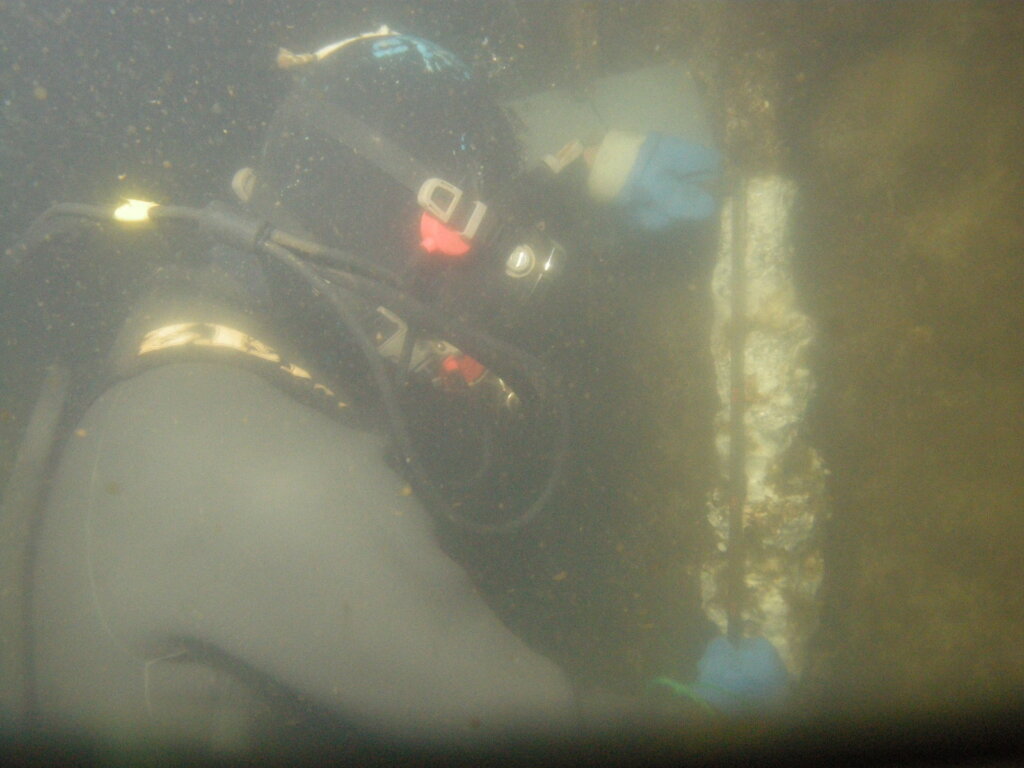 DSCN8690 - 株式会社SeaChallenge - 株式会社SeaChallenge Diver in a dark wetsuit with blue gloves exploring a murky underwater environment near a vertical wall or wreckage surface.