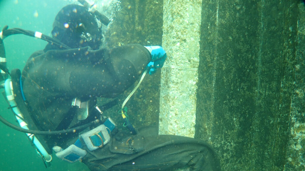 OLYMPUS DIGITAL CAMERA - 株式会社SeaChallenge Scuba diver in a black wetsuit using a tape measure to record the dimensions of a vertical concrete pillar underwater