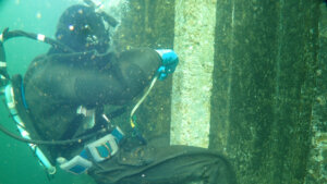 OLYMPUS DIGITAL CAMERA - 株式会社SeaChallenge Scuba diver in a black wetsuit using a tape measure to record the dimensions of a vertical concrete pillar underwater