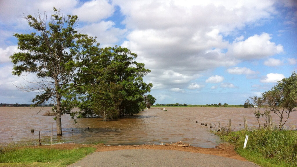 Road blocked by floodwaters with trees standing in muddy water under a blue sky and scattered clouds.