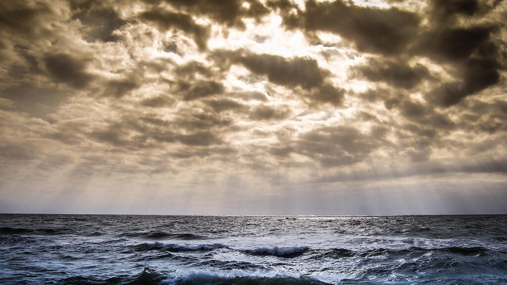 Dramatic seascape: dark clouds above a choppy ocean with sunbeams breaking through to the horizon