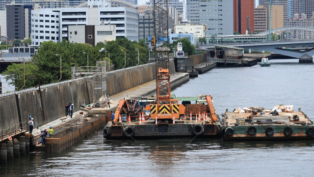 26251600_s - 株式会社SeaChallenge Construction barge with crane on a river, workers on the quay, urban skyline in the background.