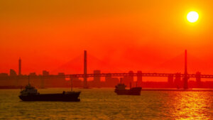 Sunset over a port with two ships on calm water and a cable-stayed bridge in the background, silhouetted against an orange sky.