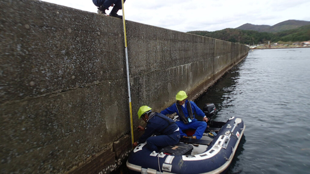 OLYMPUS DIGITAL CAMERA - 株式会社SeaChallenge Two workers in neon helmets on an inflatable boat measure depth along a seawall using a vertical measurement rod by the wall.