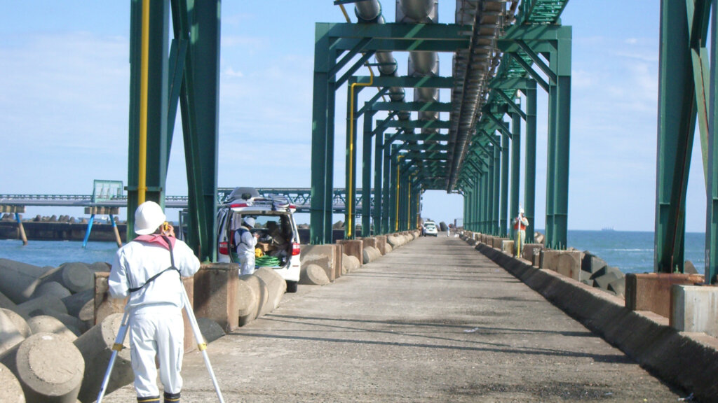 水準測量 - 株式会社SeaChallenge Surveyor in white safety gear and hard hat sets up a tripod on a concrete pier beneath a green steel gantry, with the ocean on either side and a maintenance vehicle nearby